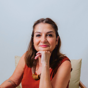 Professora e Psicóloga clínica Ana Ignez está sentada, com uma de suas mãos no queixo, com um leve sorriso. Ana é uma senhora mais velha, com algumas pequenas marcas de expressão de idade. Seu cabelo castanho escuro na altura dos ombros está penteado para trás. Ana Ignez está com um vestido laranja-avermelhado e um colar de tiras de couro sintético e madeira.