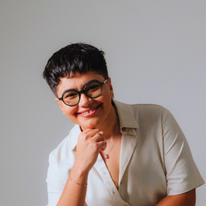 Psicóloga da clínica histórico cultural Brenna está sorrindo e veste um conjunto de camisa e calça off-white e tênis preto. Brenna tem cabelos pretos cursos, usa óculos preto e tem rosto arredondado. Diretora do Instituto Veresk de Psicologia Clínica Histórico Cultural no Brasil, teoria baseada em Vigotski.