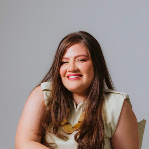 Psicóloga da clínica histórico cultural Mylene está sorrindo e usando blusa off-white com saia laranja-avermelhada. Mylene tem cabelos castanhos claros longos, rosto arredondado e lábios avermelhados. Diretora do Instituto Veresk de Psicologia Clínica Histórico Cultural no Brasil, teoria baseada em Vigotski.