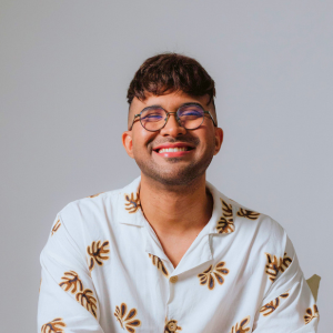 Psicólogo da clínica histórico cultural Me. Neto Oliveira está sorrindo e usando camisa off white com estampa floral marrom e calça off-white. Neto tem cabelos castanhos curtos, com uma pequena franja, barba raspada muito baixa e usa óculos redondos. Diretor do Instituto Veresk de Psicologia Clínica Histórico Cultural no Brasil, teoria baseada em Vigotski.