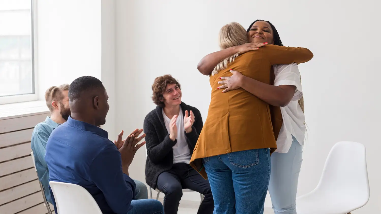 Características do terapeuta histórico-cultural. Foto de uma sala com um grupo de intervenção em que a terapeuta está abraçando  uma das pacientes. O terapeuta histórico-cultural compreende o ser humano como resultado de relações sociais, históricas e culturais. Ele se implica no processo terapêutico, não é neutro, e reconhece sua própria história e afetações. Sua escuta é intencional, voltada ao desenvolvimento do paciente, promovendo novas formas de compreender e lidar com o sofrimento. Atua de forma crítica, afetiva e ética, entendendo o sofrimento em sua dimensão social e política, comprometido com práticas antidiscriminatórias e com a promoção da dignidade humana.